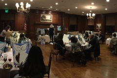 Panoramic view of people grouped at circular tables, all painting the same image as the demonstrator in the middle.
