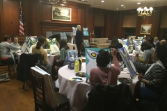 People seated at tables, all painting the same colorful creature while one person demonstrates.